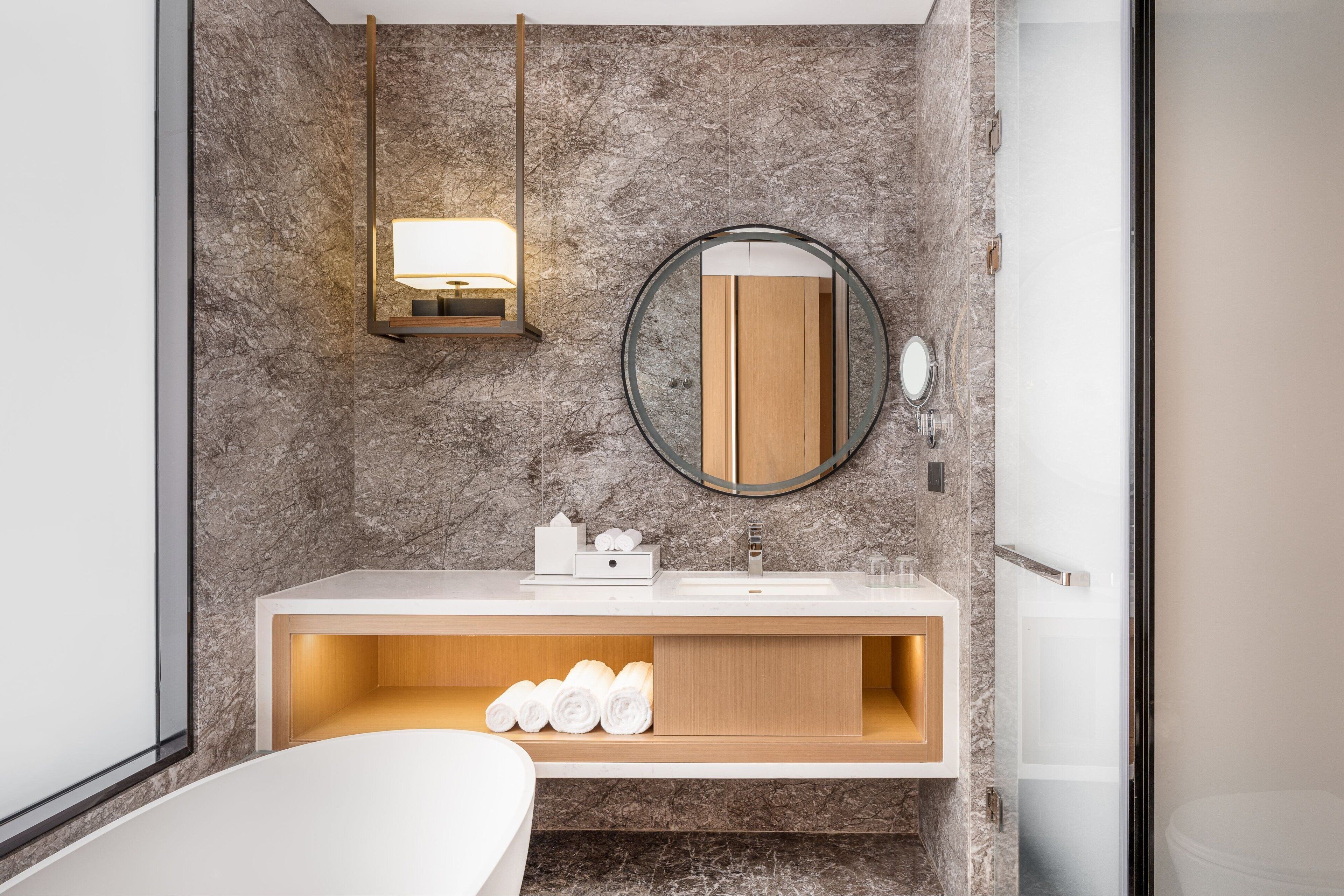 A modern bathroom with a marble wall, a round mirror, a built-in sink, and a shelf with towels.