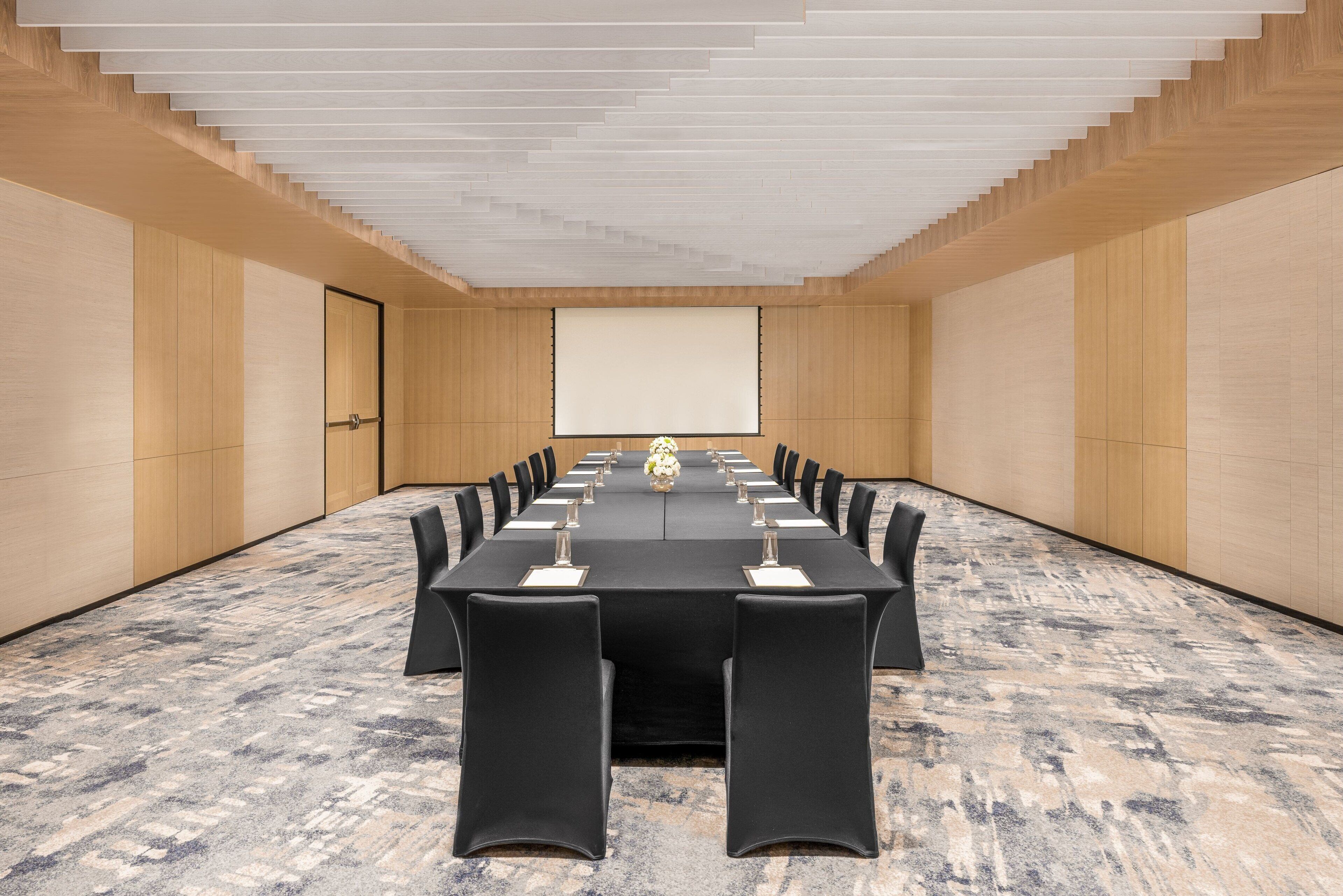 Una sala de conferencias con una larga mesa, sillas, una pantalla de proyector y piso alfombrado.
