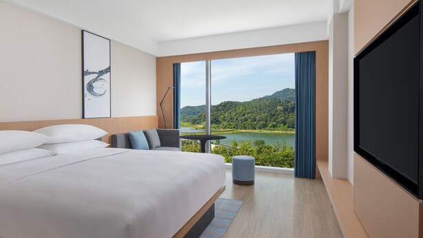 Una habitación de hotel moderna con una cama grande, un escritorio y un televisor. La habitación tiene un ventanal con vistas a un lago y montañas.