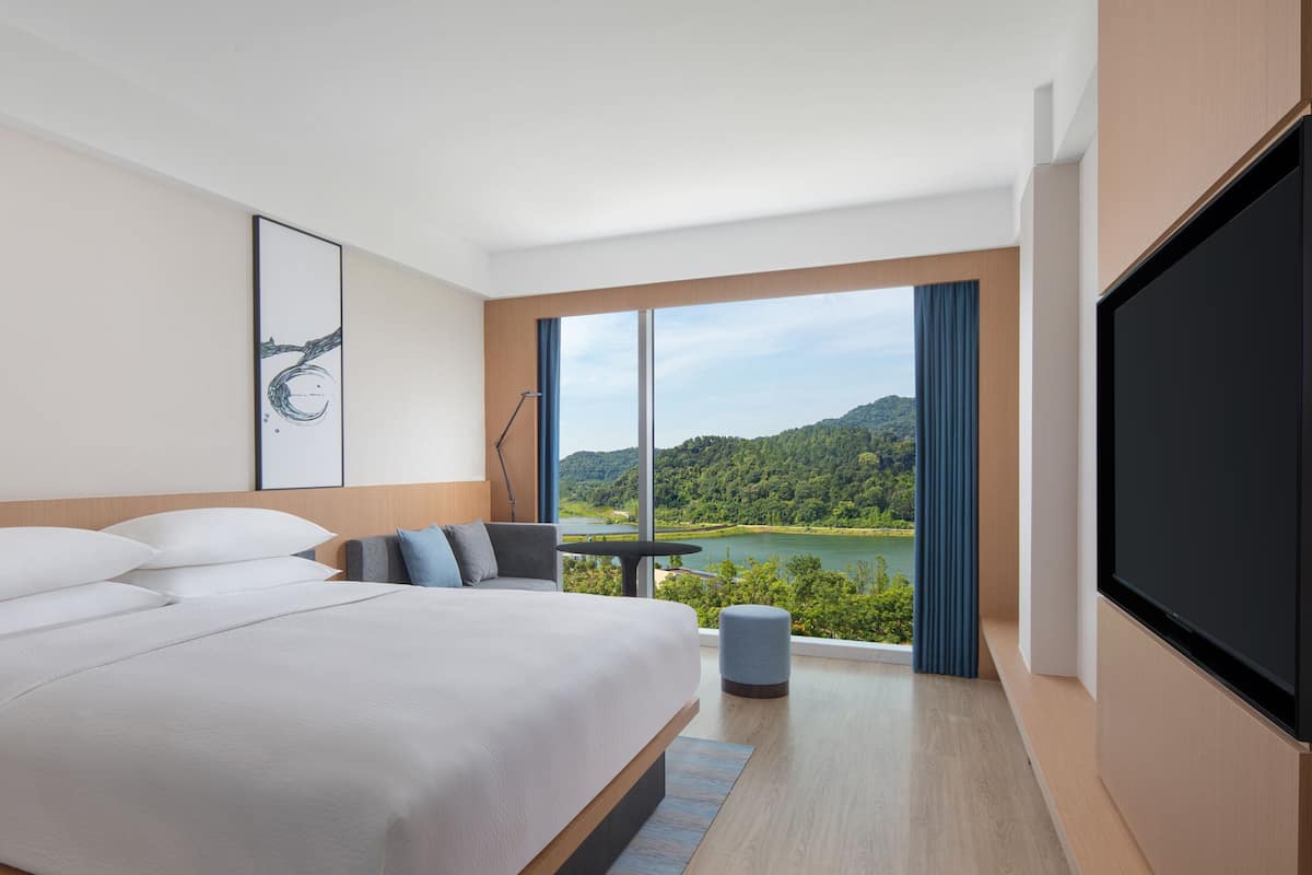 A modern hotel room with a large bed, a desk, and a TV. The room has a large window with a view of a lake and mountains.