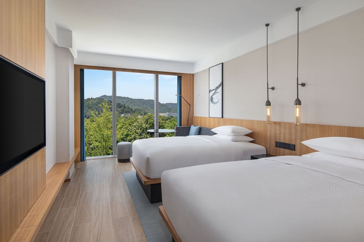 A modern hotel room with two beds, a large TV, and a view of greenery.