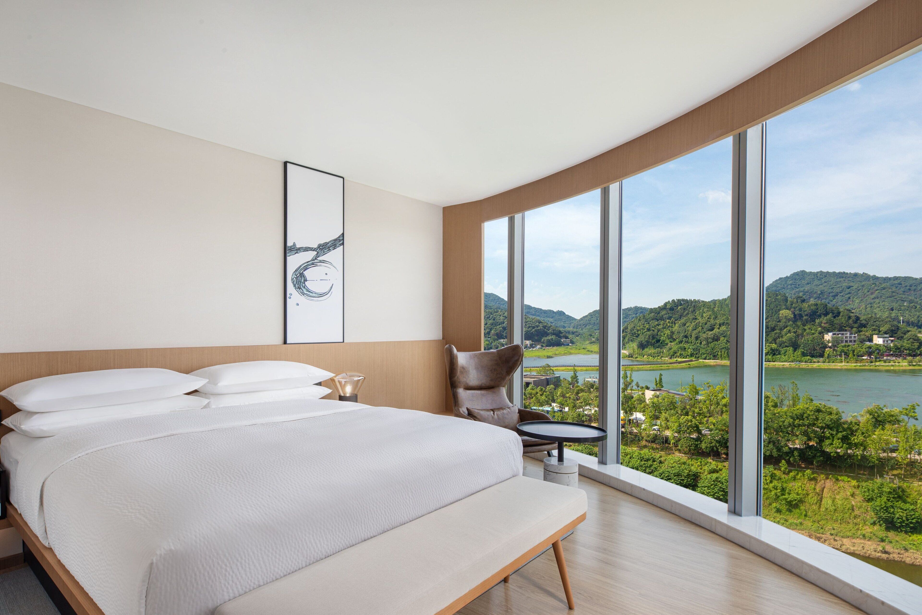A modern hotel room with a large bed, a chair, a small table, and a large window offering a view of a lake and hills.
