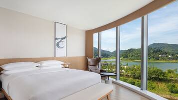 A modern hotel room with a large bed, a chair, a small table, and a large window offering a view of a lake and hills.
