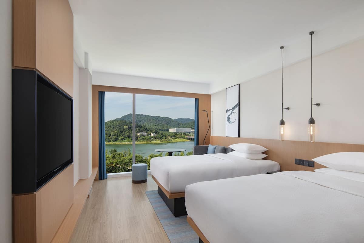 A modern hotel room with two beds, a flat-screen TV, and a large window offering a scenic view of a lake and hills.