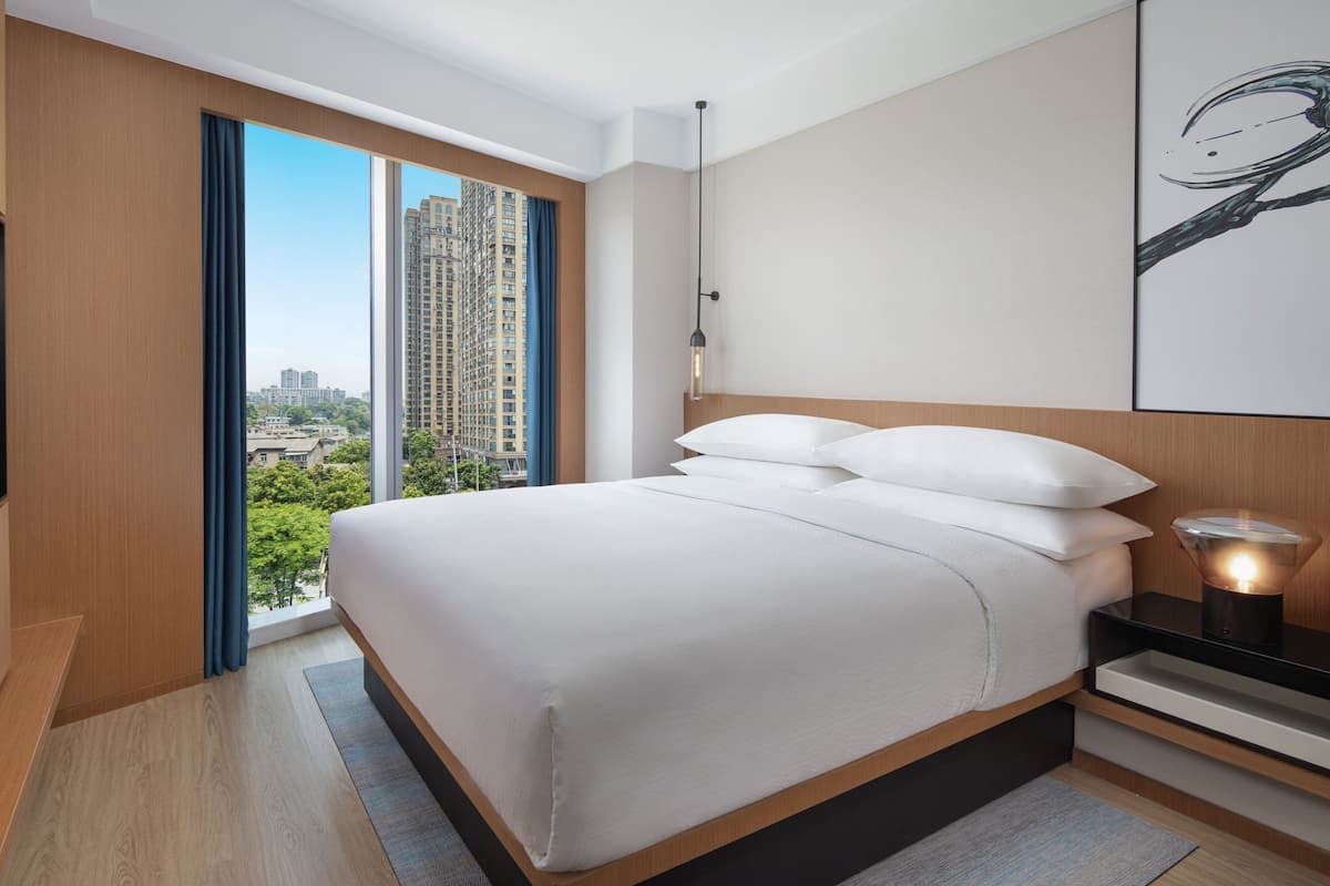 A modern hotel room with a large bed, a nightstand with a lamp, and a view of a cityscape through a large window.