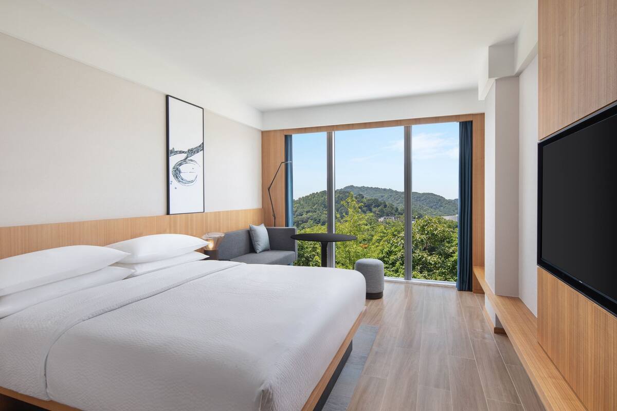 A modern hotel room with a large bed, a seating area, a flat-screen TV, and a view of the outdoors.