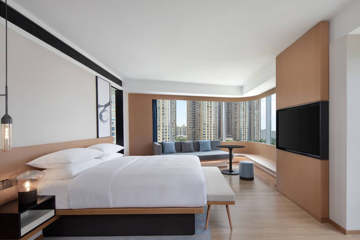 A modern hotel room with a large bed, a seating area with a sofa and a small table, a flat-screen TV, and a view of high-rise buildings.