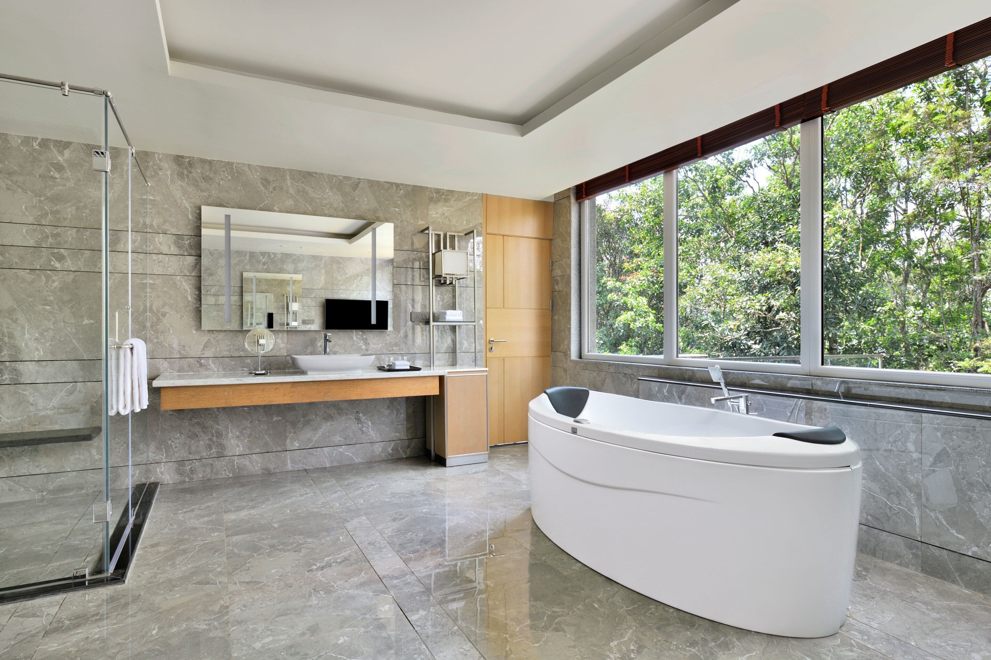 A modern bathroom with a glass shower, a large bathtub, a vanity with a sink, and a large window with a view of trees.