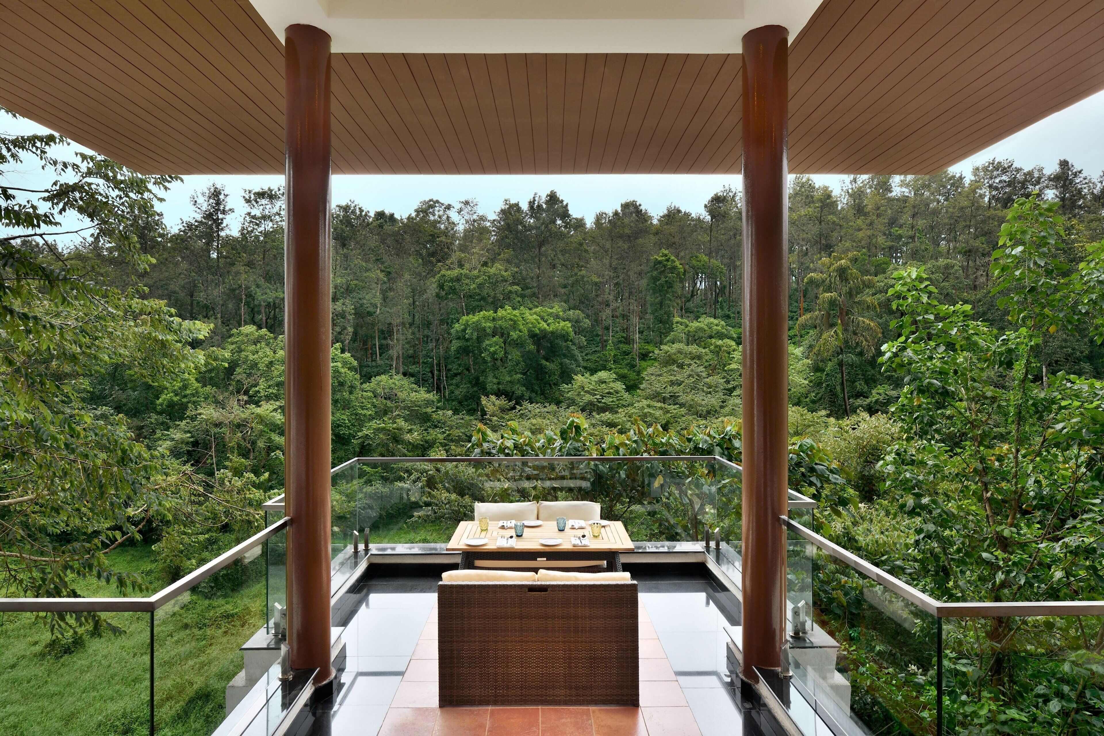 A balcony with a dining table and chairs, surrounded by glass railings, overlooking a lush forest.