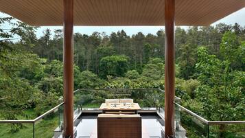 A balcony with a dining table and chairs, surrounded by glass railings, overlooking a lush forest.