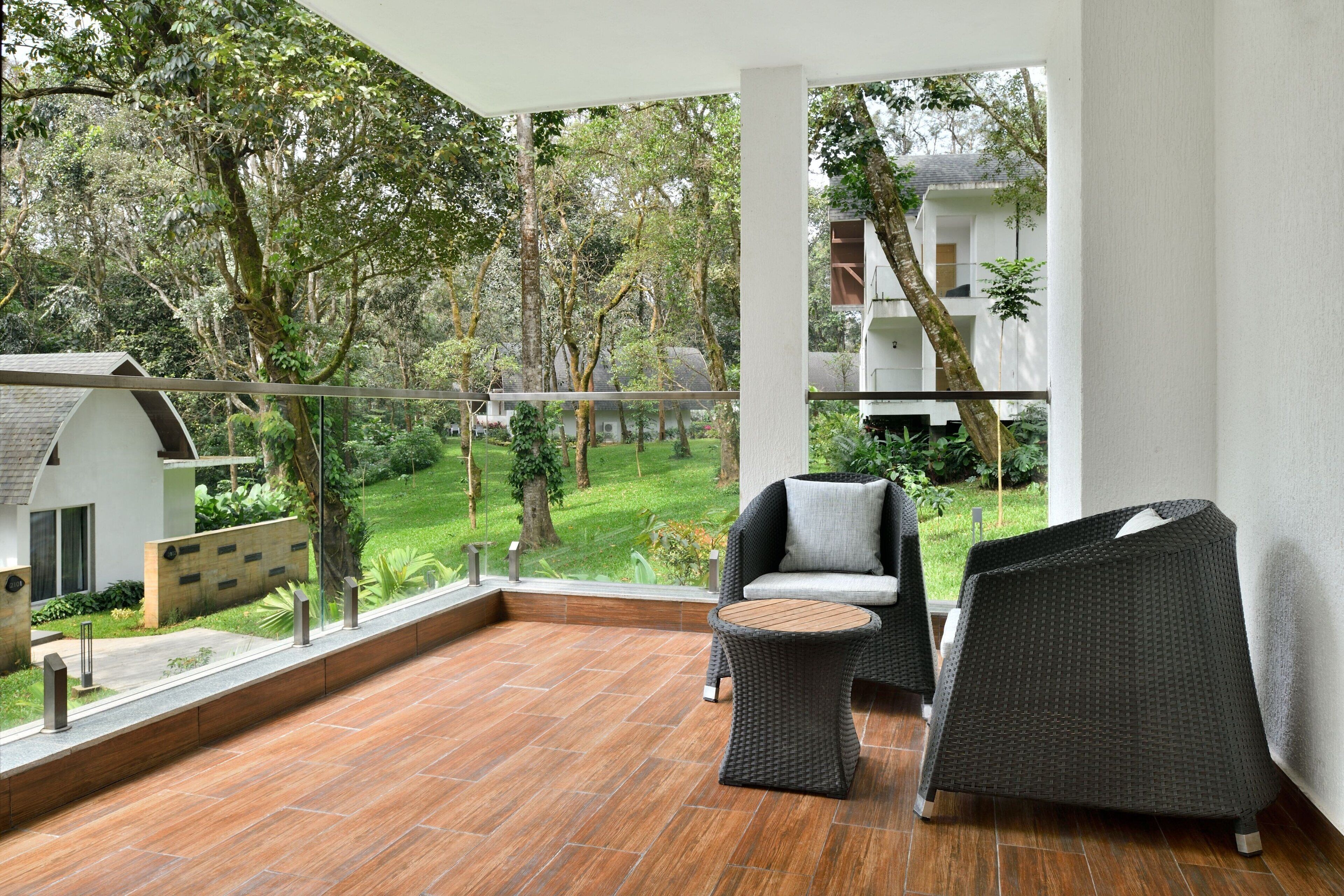 A balcony with a wicker chair and a small table, overlooking a lush garden and a white building.