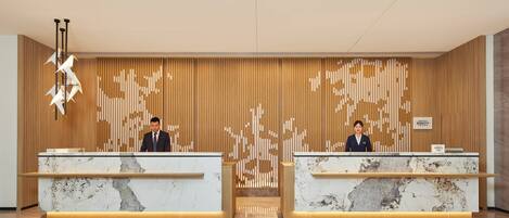A hotel reception area with two receptionists, wooden paneling, marble reception desks, and a modern light fixture.