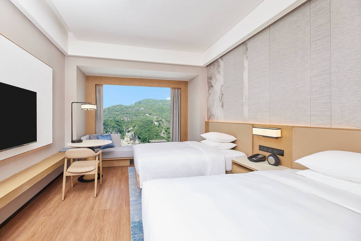 Une chambre d’hôtel avec deux lits, un bureau avec une chaise, une télévision et une fenêtre donnant sur la montagne.