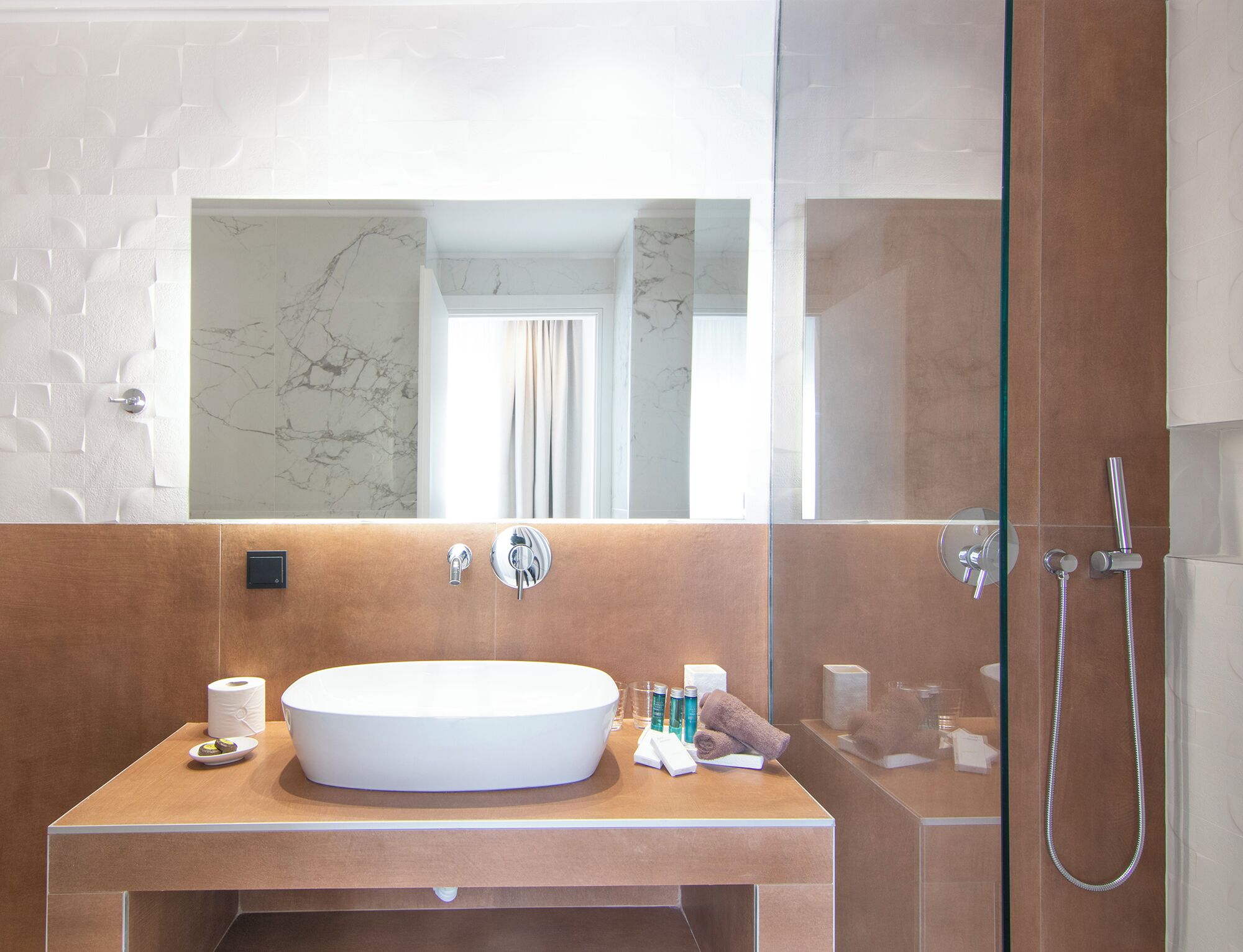 A bathroom with a marble wall, a large mirror, a round sink, and a shower area.