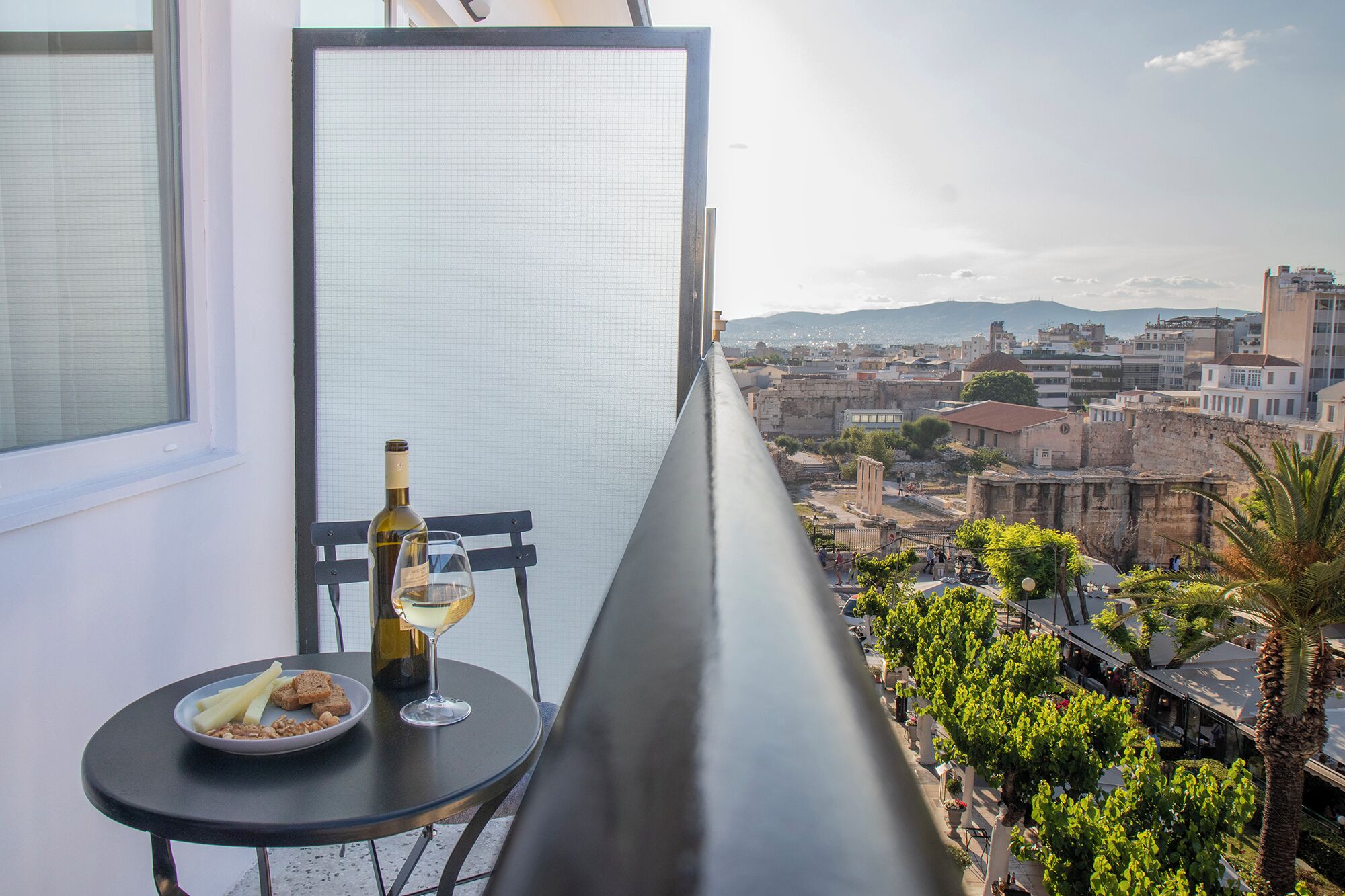 Un balcón con una mesa dispuesta para dos personas, una botella de vino y un plato de comida. Se observa una vista urbana con edificios y una cadena montañosa distante.