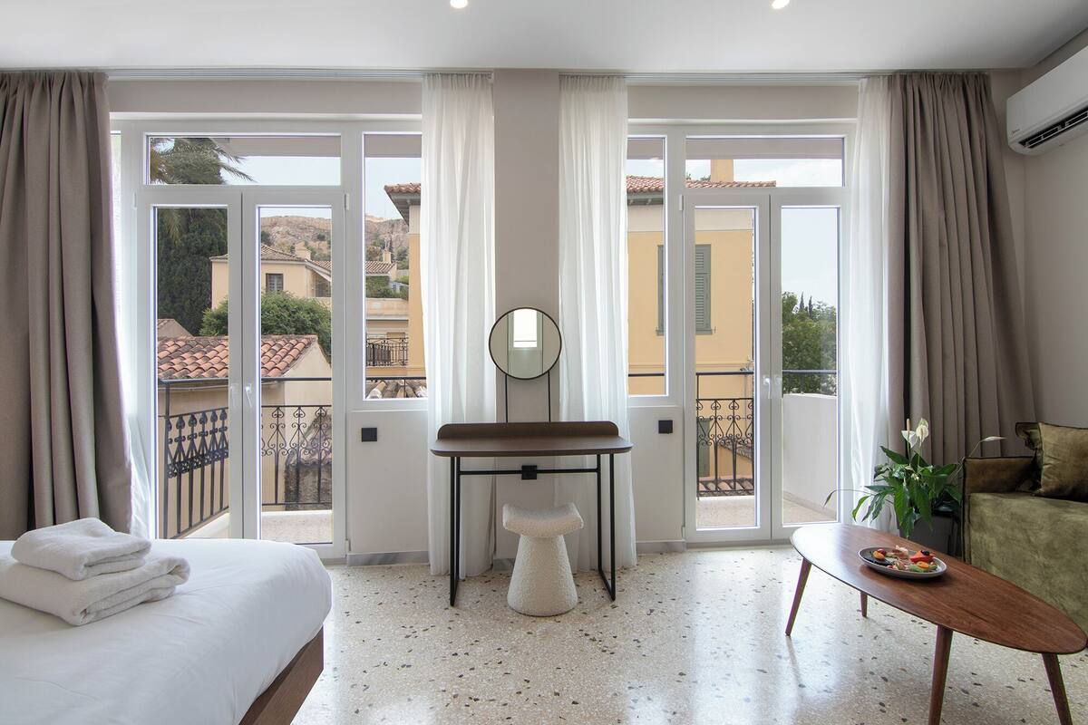 A hotel room with a bed, a small table with a mirror, a sofa, and a balcony with a view of buildings.