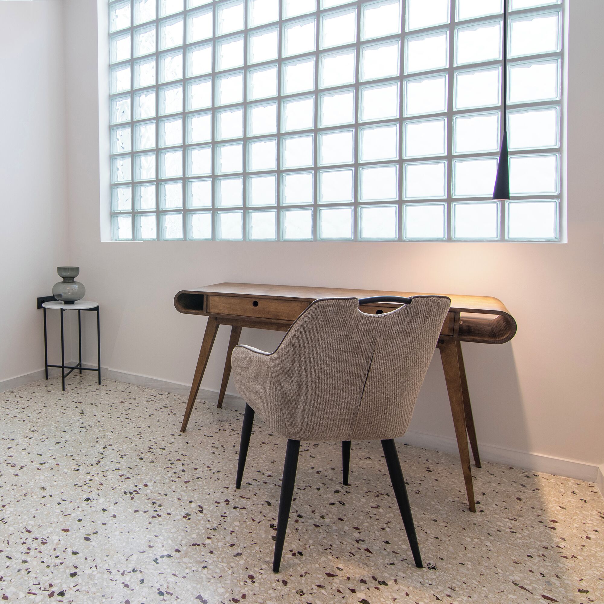 Quarto com uma janela ampla, uma escrivaninha de madeira, uma cadeira e uma pequena mesa com um vaso.