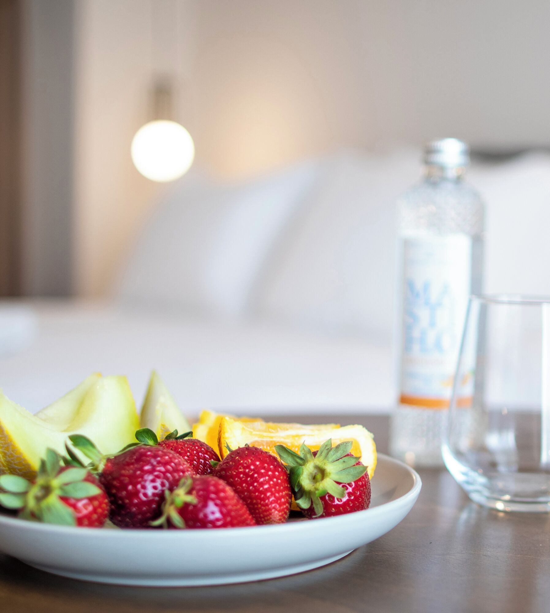 A plate of fresh fruit including strawberries and oranges, a glass of water, and a bottle of water on a wooden table in a hotel room.