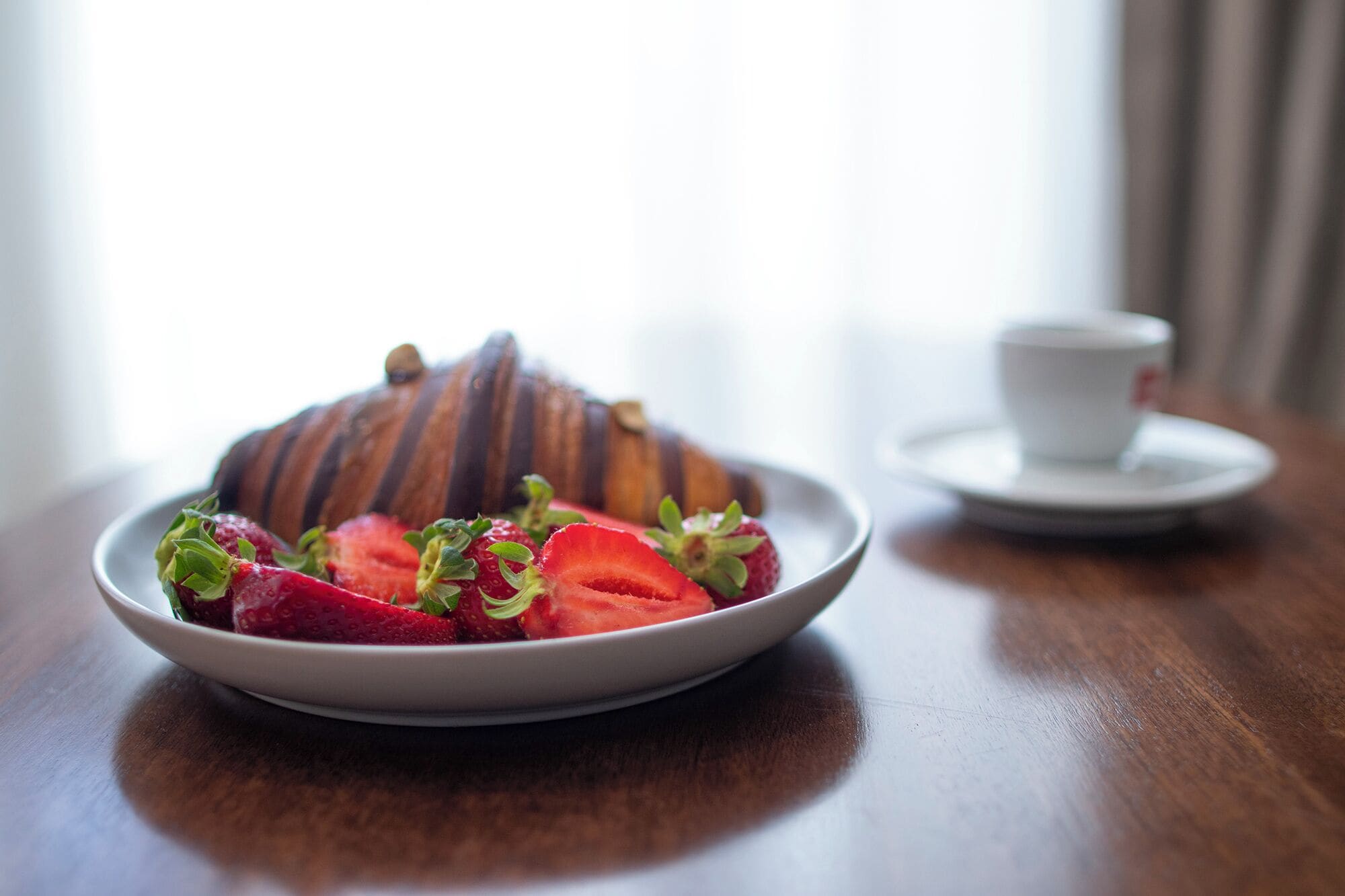 A plate of sliced strawberries and a croissant on a wooden table with a white cup and saucer in the background.