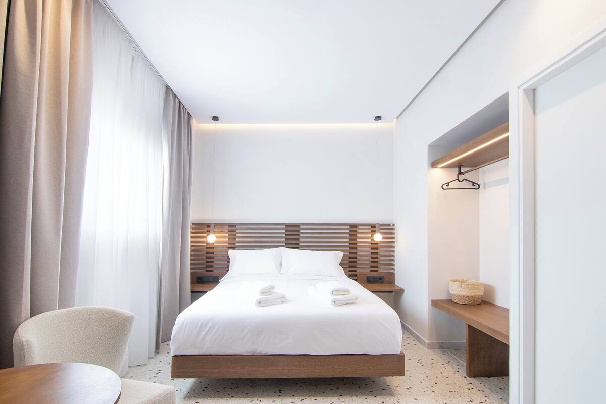 A modern hotel room with a large bed, a wooden headboard, a bedside table, a chair, and a bench.