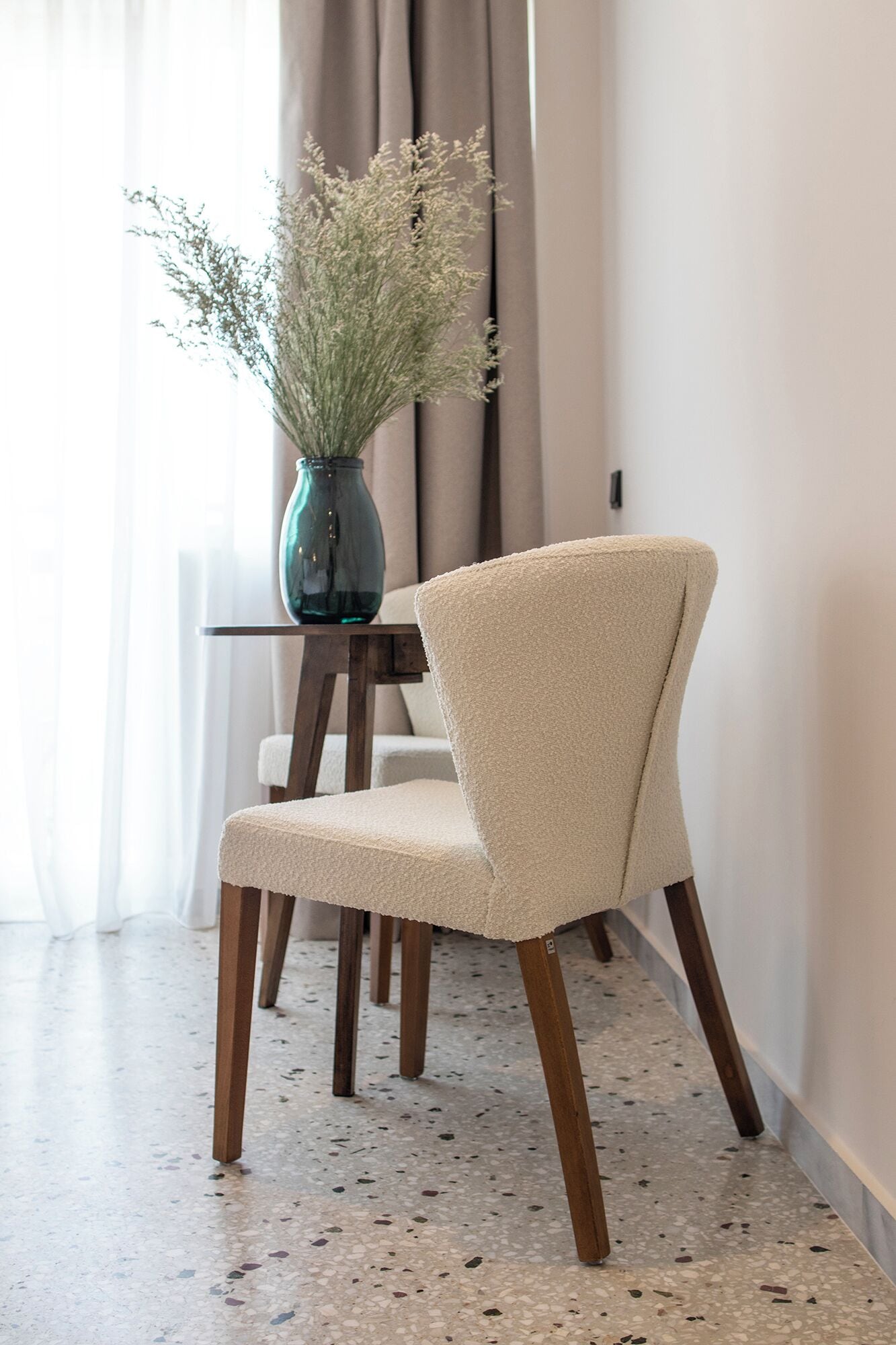 A chair with a textured fabric seat and backrest, wooden legs, and a vase with dried flowers on a small table.