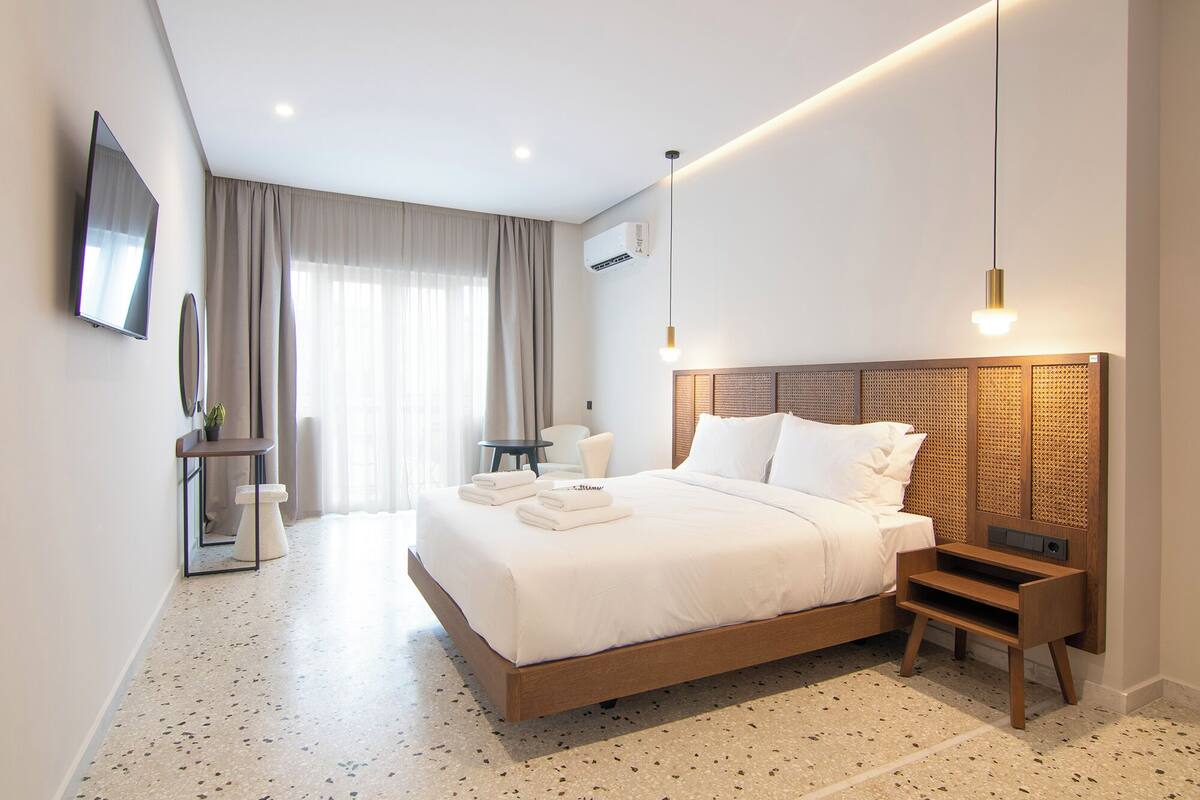 A modern hotel room with a large bed, a bedside table, a small table with a chair, a TV mounted on the wall, and a window with curtains.
