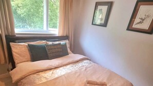 Una cama bien hecha con un cojín estampado, una ventana con cortinas y dos cuadros en la pared.