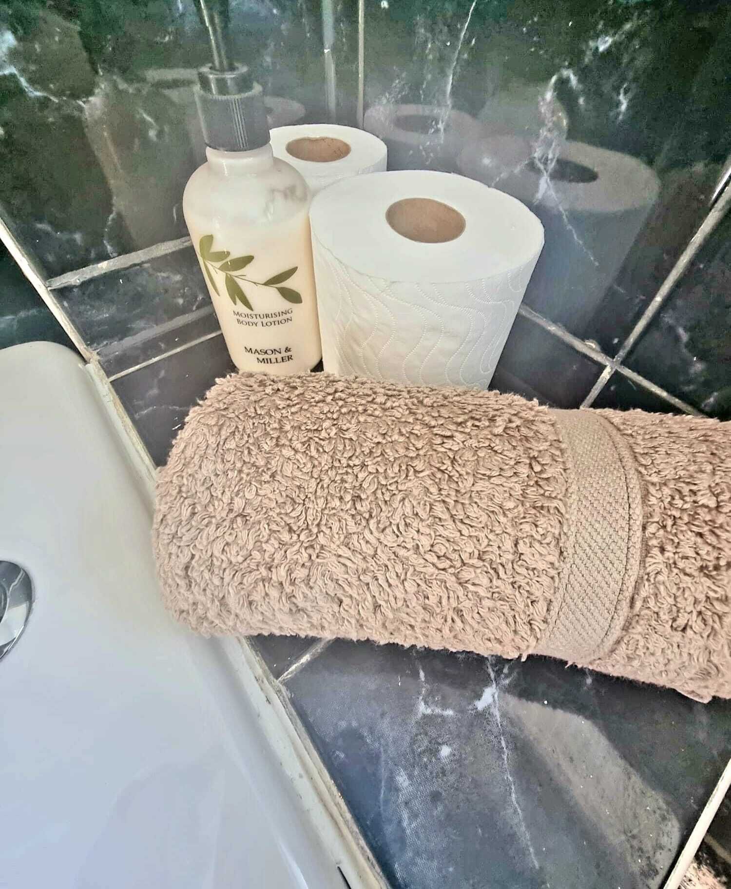 A bathroom with a beige towel, a bottle of moisturizing body lotion, and two rolls of toilet paper on a tiled floor.