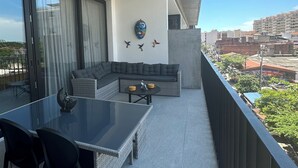 A balcony with a glass table and chairs, a grey sofa, and a decorative wall piece.