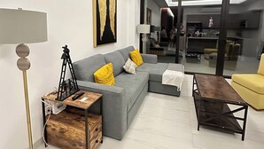 A modern living room with a grey sectional sofa, a wooden coffee table, a floor lamp, and a large window offering a view of the interior.