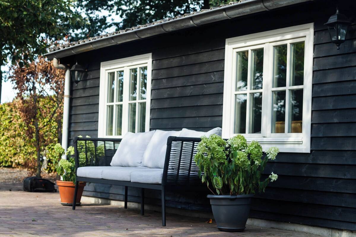 A black wooden building with two white-framed windows, a black bench with white cushions, and potted plants.