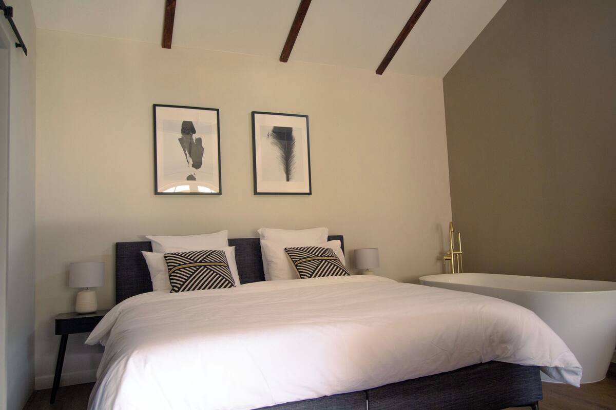 A bedroom with a large bed, two framed pictures on the wall, a side table with a lamp, and a freestanding bathtub.