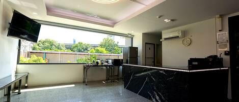 A modern hotel lobby with a marble reception desk, a flat-screen TV mounted on the wall, and a large window offering a view of the outdoors.
