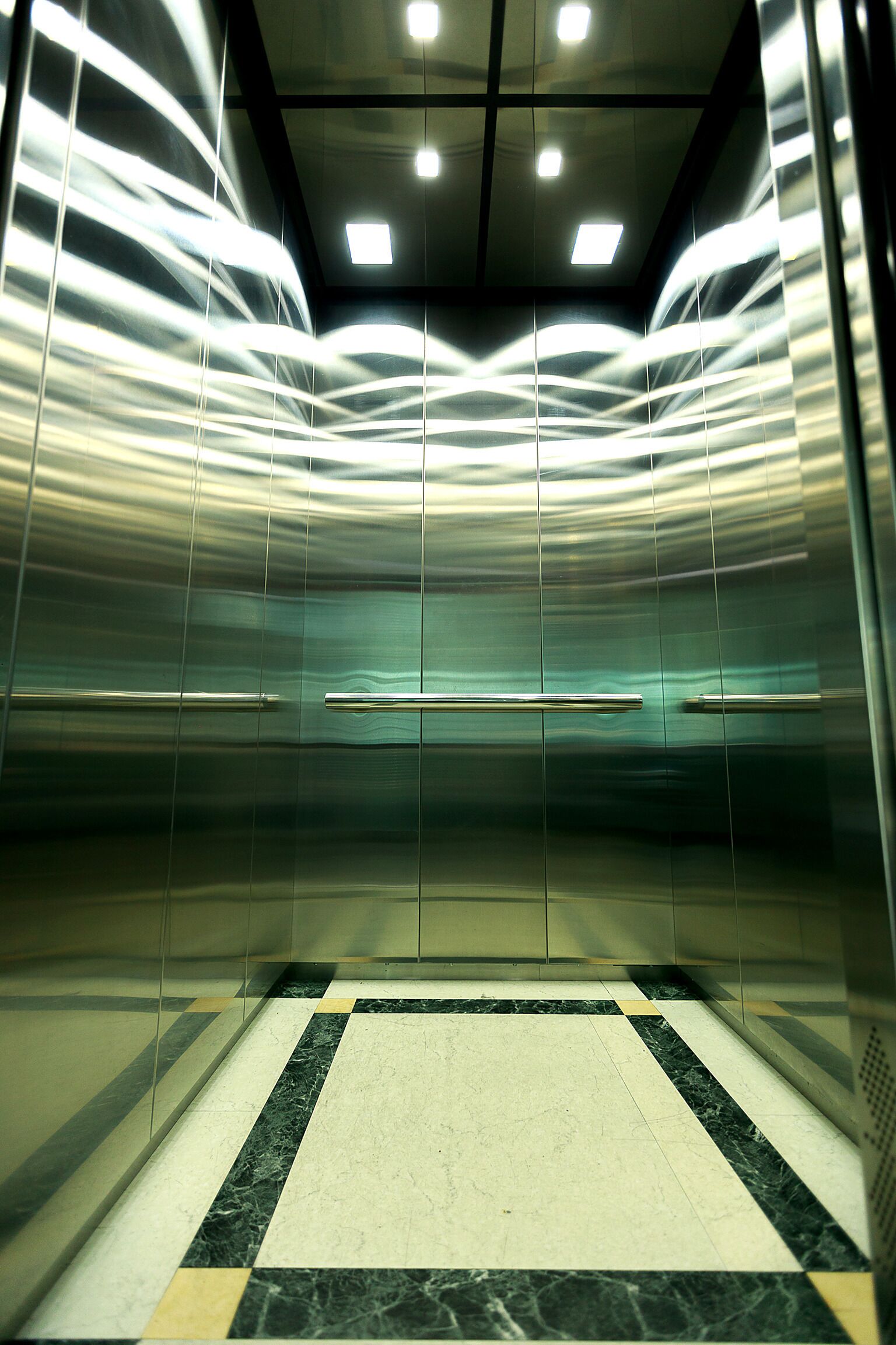 An interior of an elevator with metallic walls and a marble floor.