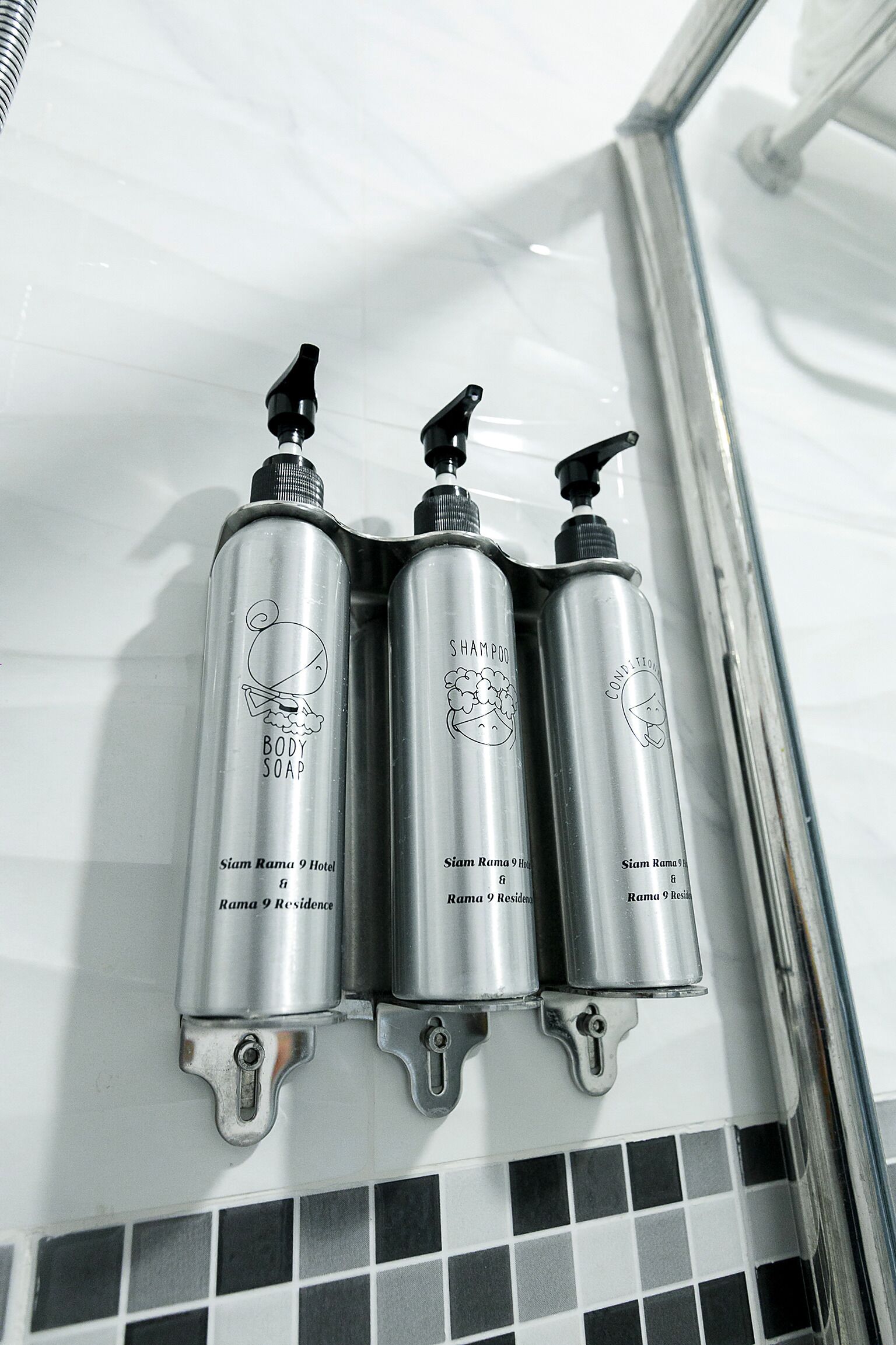Three metallic bottles with pump dispensers, labeled 'Shampoo Body Soap,' mounted on a tiled wall.