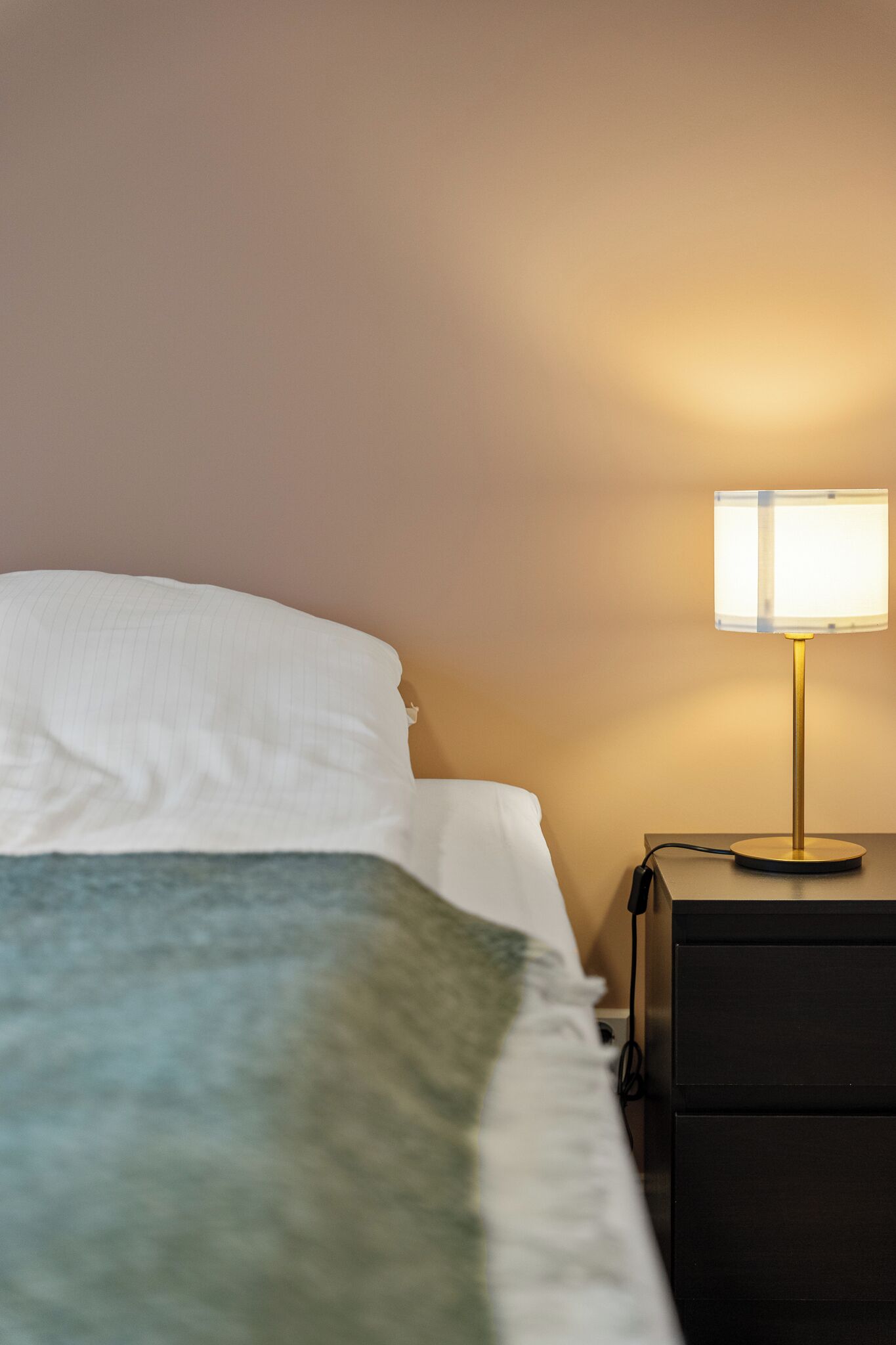 Una cama bien hecha con una almohada blanca y una manta verde, una mesita de noche con una lámpara y un tocador negro.