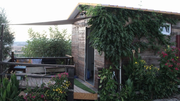 Ein rustikales Holzhaus mit einer Holzterrasse, einem kleinen Garten mit verschiedenen Pflanzen und Blumen sowie einer Aussicht auf die Landschaft im Hintergrund.