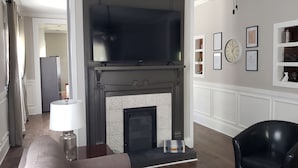 Een woonkamer met een open haard, een zwarte leren fauteuil, een lamp en lijstjes met foto's aan de muur.