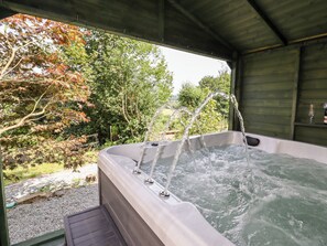 A hot tub with multiple jets, surrounded by a wooden deck and a view of trees and greenery. - Trem Y Don (Conwy)