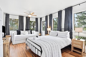 A bedroom with a bed, a white sofa, a nightstand, a ceiling fan, and large windows with curtains.