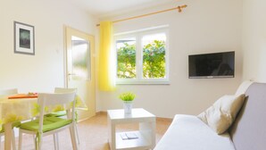 Un salon lumineux avec un canapé, un téléviseur fixé au mur, un coin repas avec des chaises et une table, et une fenêtre donnant sur un paysage verdoyant.
