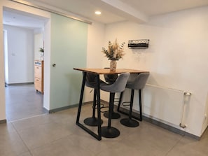 Un comedor con una mesa de madera y dos taburetes altos, un jarrón con flores secas y un altavoz en la pared.