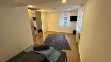 A modern, compact living space with a bed, a small seating area, a flat-screen TV, and a window with curtains.