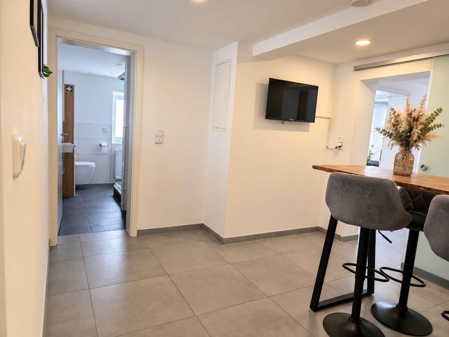 A modern kitchen with a flat-screen TV mounted above a wooden countertop, two bar stools, and a visible bathroom with a toilet and sink.
