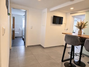 Una cocina moderna con una televisión de pantalla plana sobre una encimera de madera, dos taburetes de bar y un baño a la vista con inodoro y lavabo.