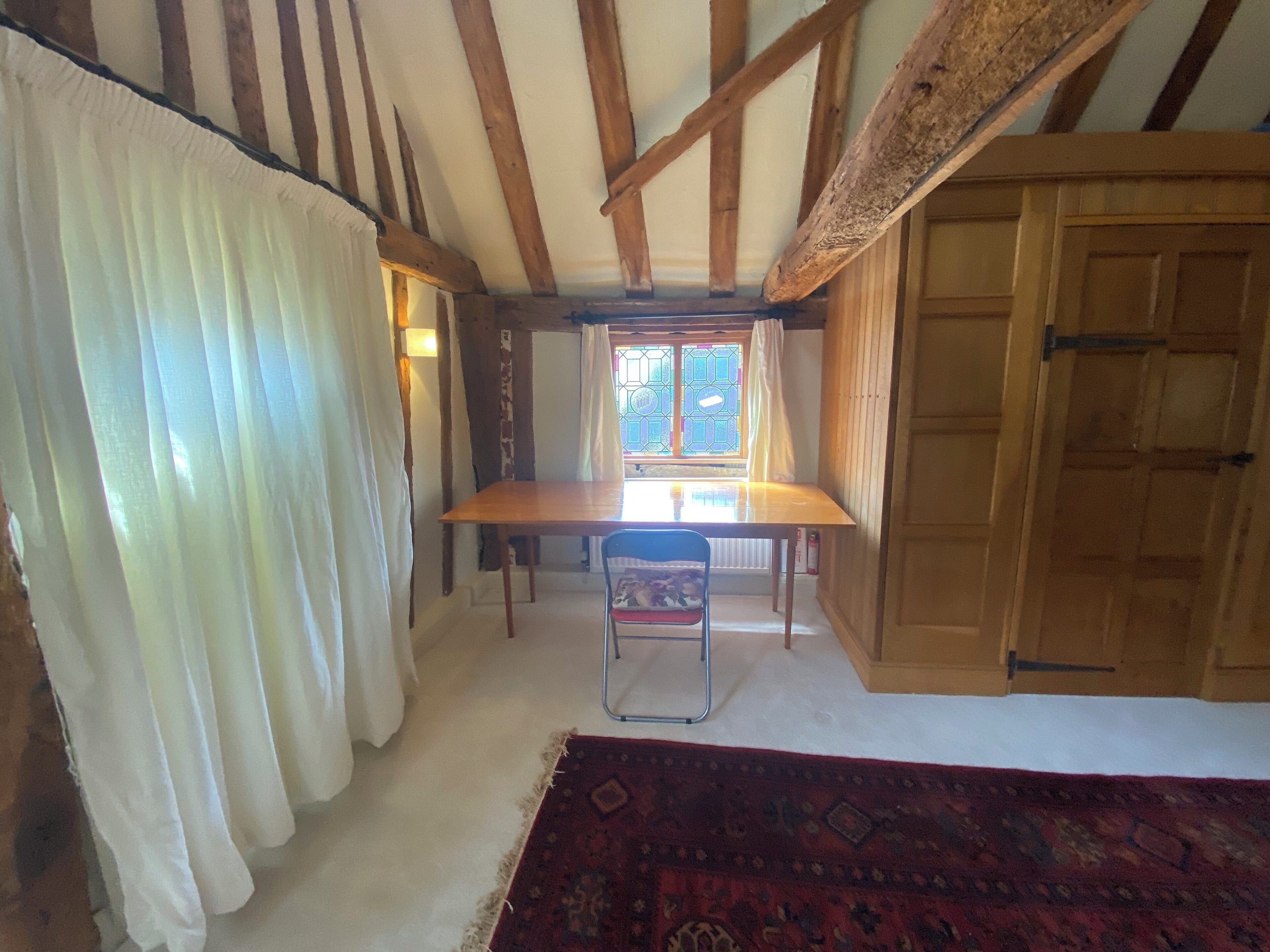 A room with a wooden ceiling, a table, a chair, a window with curtains, and a patterned rug.