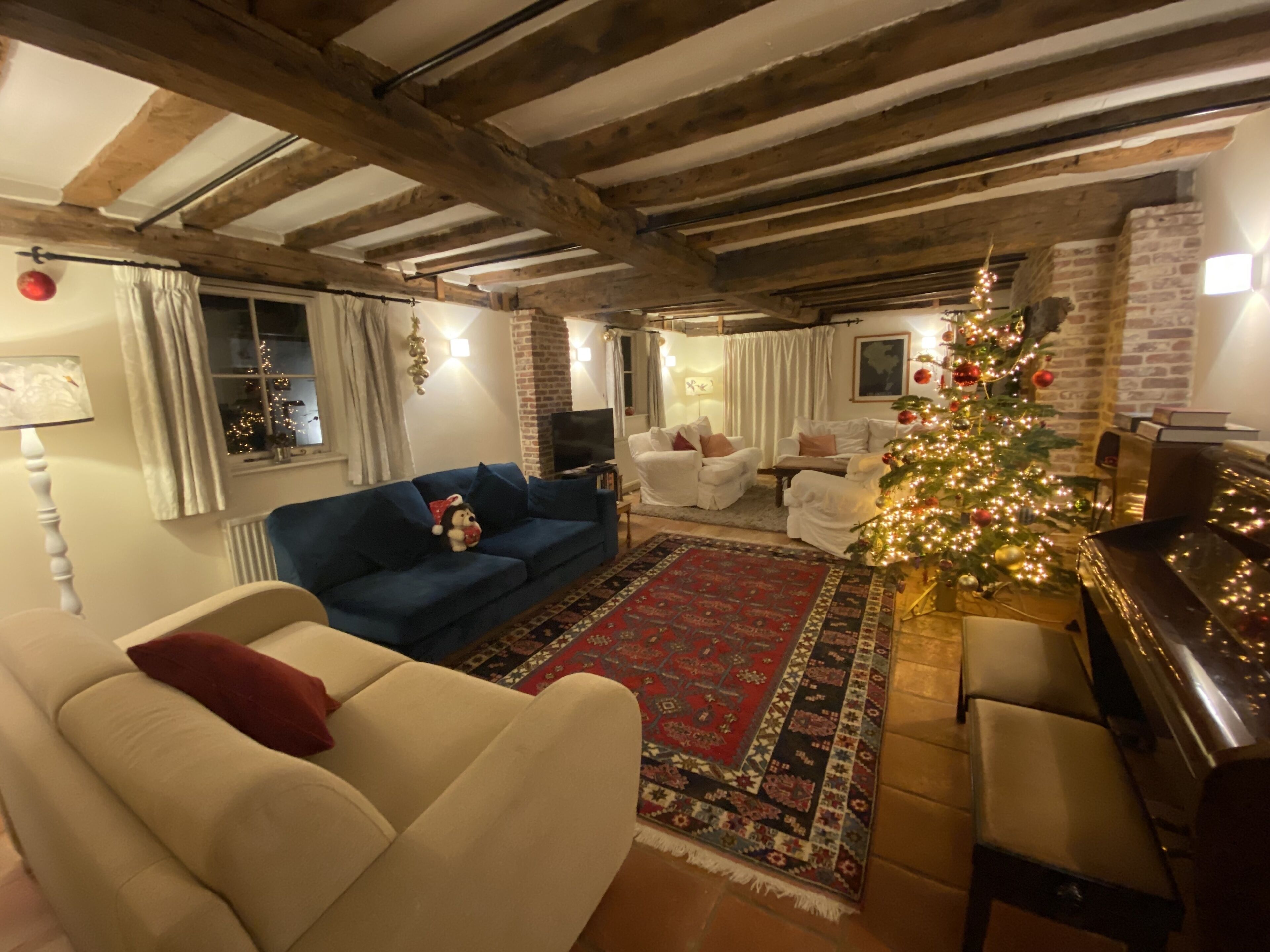A cozy living room with a blue sofa, white armchairs, a red and black patterned rug, a Christmas tree, and a piano.