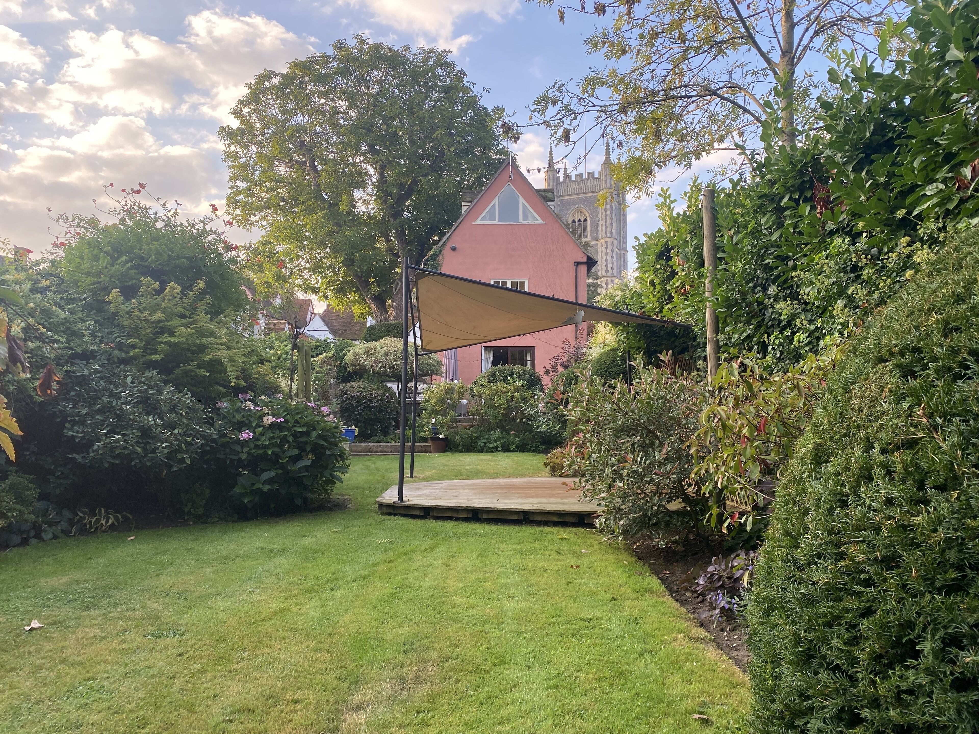 A garden with a wooden deck, a red building with a tower, and a large tree.