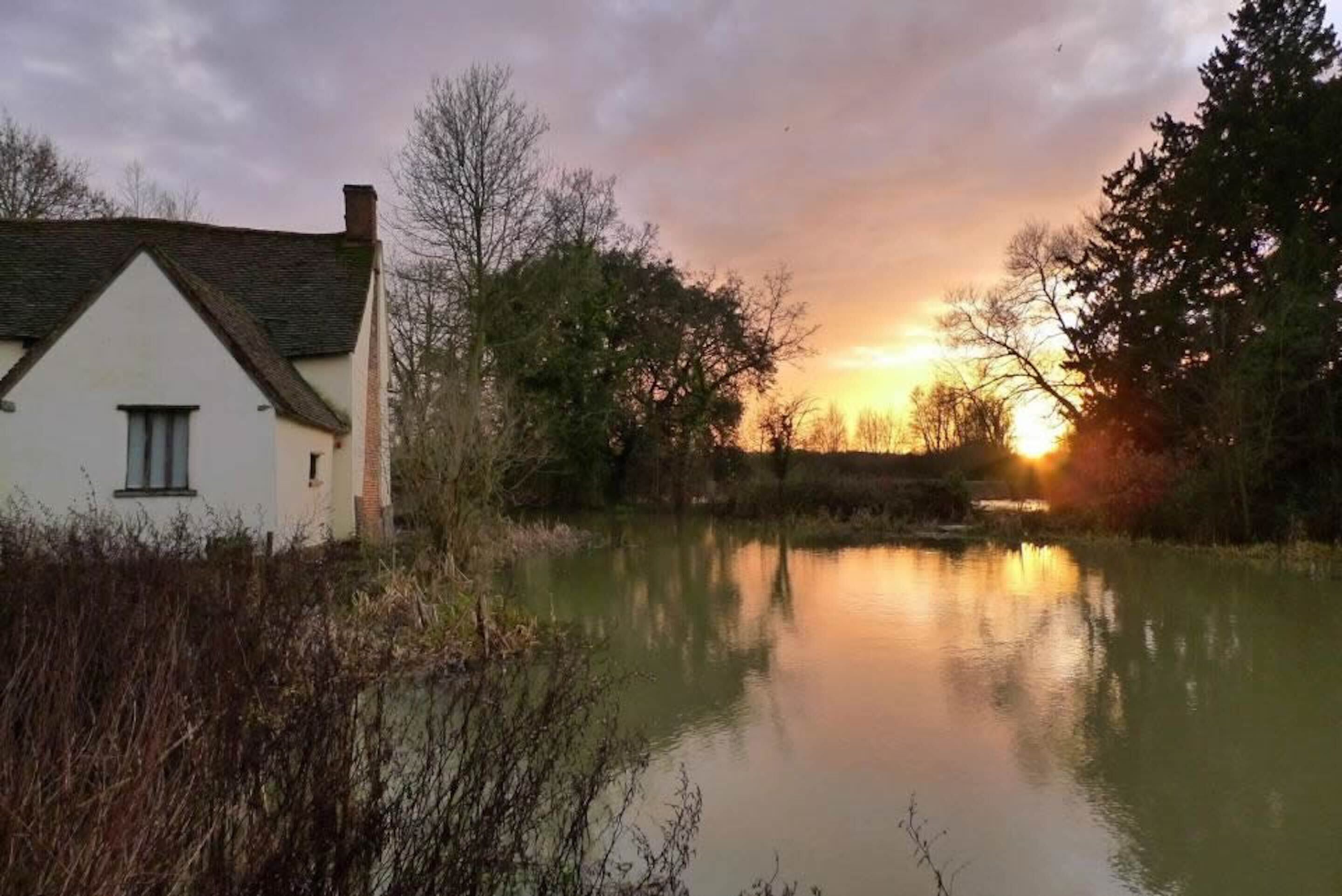 Una casa bianca con tetto a capanna vicino a una superficie d'acqua calma che riflette il tramonto.