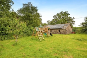 A rustic stone building with a sloped roof, a wooden swing set, and a ladder in a grassy yard. - 2 Bed in Torrington (oc-p27344) (Langtree)